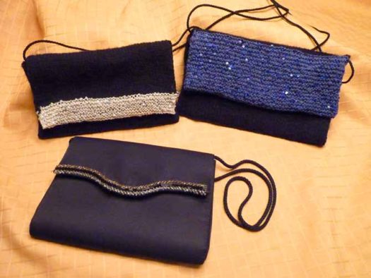 The two knitted bags, small one on the left and large, blue on on the right, propped up against a gold cloth, with a black fabric bag that has a beaded edge on the flap laying in the foreground