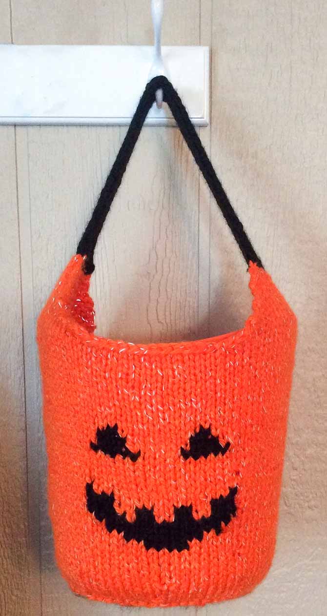 An orange knitted bag with a jack-o-lantern face and handle made in black, hanging from a hook.