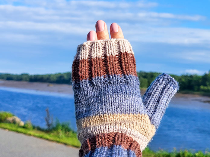 A left hand wearing blue and brown striped open finger mitts knit from UNIVERSAL YARN Deluxe Stripes, color Timber, against a background of a walking trail along a river.