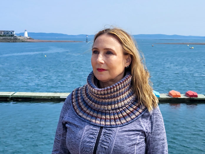 Michelle Porter is wearing a blue and brown striped cowl, knit from UNIVERSAL YARN Deluxe Stripes, color Timber, with a lighthouse, ocean, and boats on a dock in the background 