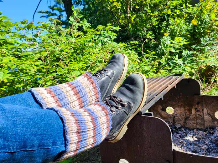 A picture of legs in denim jeans and black hiking boots wearing the blue and brown striped boot cuffs knit from UNIVERSAL YARN Deluxe Stripes, color Timber, feet propped up onto a metal fireplace. 