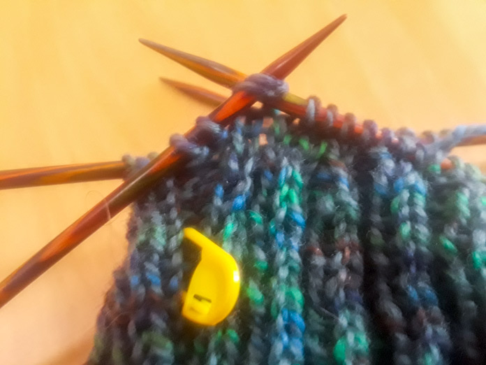 Working the crown decreases of the Gallinule Hat in Fibra Natura Cobblestone Yarn. Yarn is the colorway “Mallard,” a marled blue, green and gray. The knitting is on a Knit Picks Wooden Double Pointed Needles set, with a UNIQUE Locking Stitch marker clipped into the knitting. 