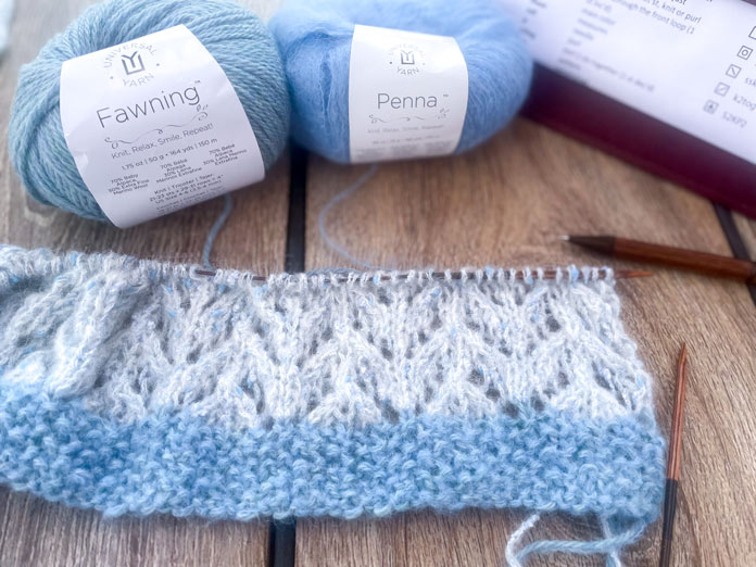 A knitted portion of Lace Tipped Cardigan using UNIVERSAL YARN Thula in the Delphinium colorway, one ball of UNIVERSAL YARN Fawning in the Moonstone colorway, one ball of UNIVERSAL YARN Penna in the Clouded colorway, and a pen on a table.