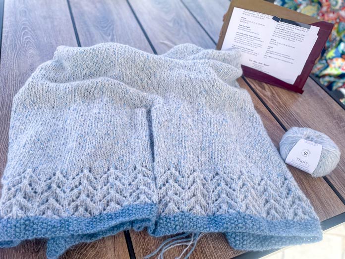 A knitted portion of Lace Tipped Cardigan, one ball of UNIVERSAL YARN Thula in the Delphinium colorway, and Knitpro Ginger Special Deluxe Interchangeable Needle Set on a table.