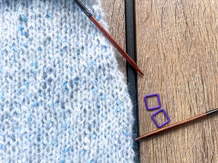 A knitted portion of Lace Tipped Cardigan using UNIVERSAL YARN Thula in Delphinium, two knitting needles and two markers from the Knitpro Ginger Special Deluxe Interchangeable Needle Set on a table.