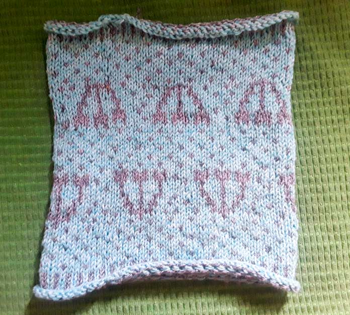 The Holland Cowl lying flat. It's a colorwork cowl knit in two colors of Mystical Marl yarns with a centered tulip motif. The bottom and top edges of the cowl are curling.