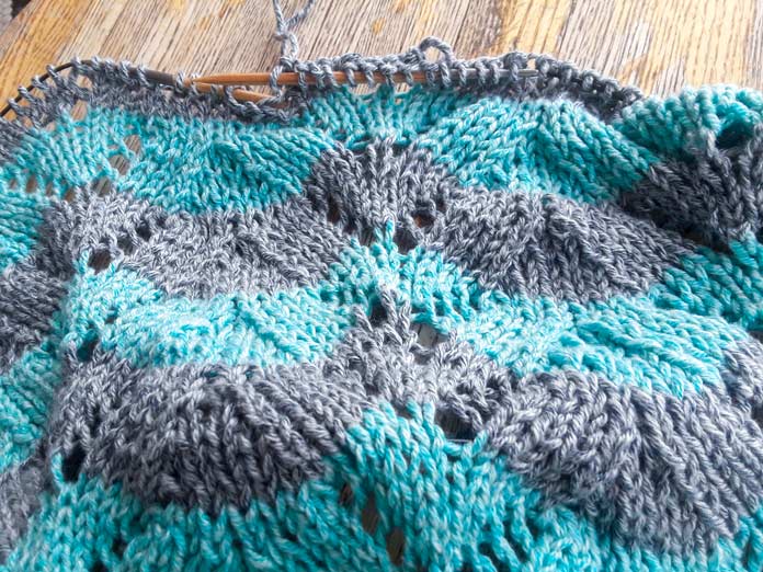 The Sunshine Throw Worsted blanket on the needles in two colors of Mystical Marl Yarn. The stripes of the colors have been pulled into a sideways zigzag pattern. 