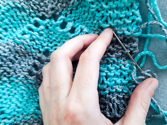 The wrong side of the Sunshine Throw with a number ends loose on one side. Someone is weaving those ends into the wrong side of the garter stitch edging. 