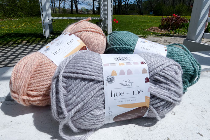 Three balls of yarn with a white ball band rest on a worn white bench with an arbor and trees in the background. A soft gray yarn (haze) is in the foreground with rosewater (a soft peach) behind it on the left, and agave, a soft medium green almost the color of blue spruce, on the right.