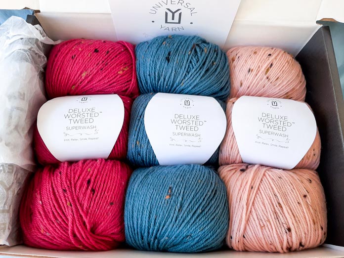 9 balls of yarn; 3 in red, 3 in blue, 3 in a light putty color packed in a box; Universal Yarn Deluxe Worsted Tweed Superwash