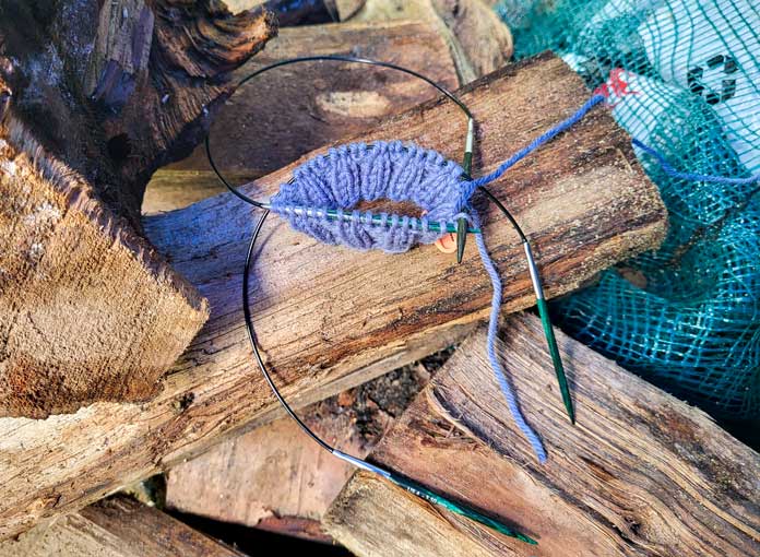 Resting on some split campfire wood is a blue mitt cuff, worked in UNIVERSAL YARN Deluxe Stripes, color Timber, knit on 2 KNITTER’S PRIDE Dreamz size circular needles.