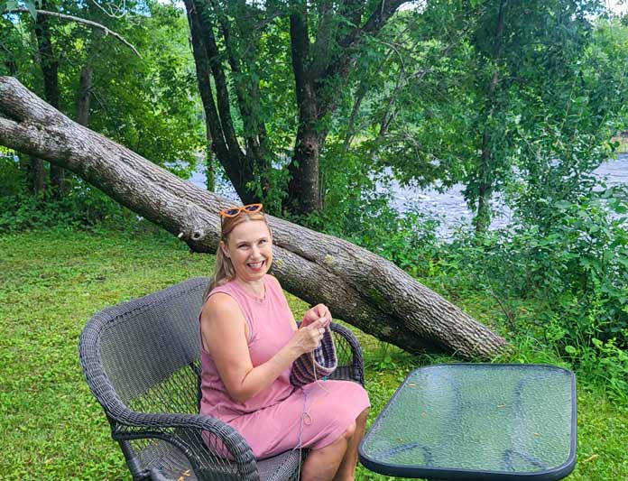 Michelle Porter is seated on a wicker garden bench as she knits a blue and brown striped cowl, worked in UNIVERSAL YARN Deluxe Stripes, color Timber on KNITTER’S PRIDE Dreamz circular needles, with a scenic river in the background.