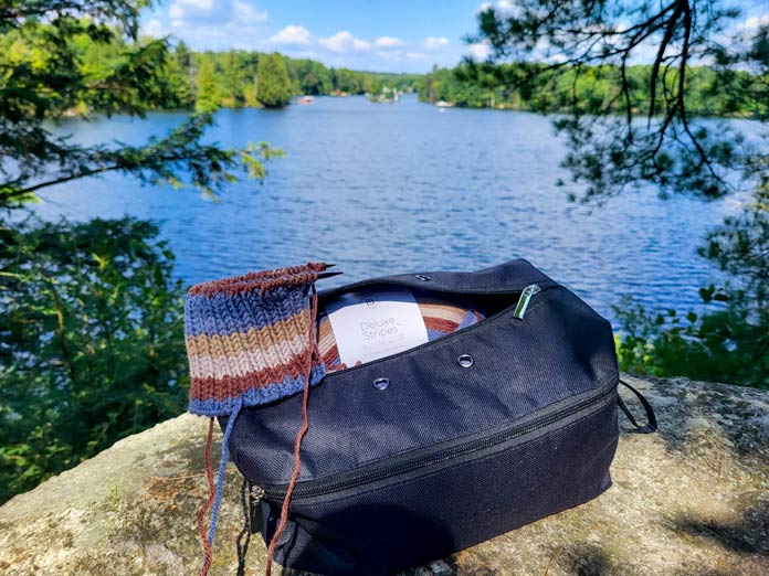 A view of the lake from the rock cliff, with the open black VIVACE Knitting bag showing a ball of UNIVERSAL YARN Deluxe Stripes, color Timber, and a blue and brown striped boot cuff being knit on KNITTER’S PRIDE Dreamz circular needles.