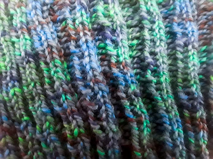 This is a close-up of the Gallinule Hat knit two, purl two ribbing, done in Fibra Natura Cobblestone yarn in Mallard. The stitches are marled blue, green, and gray. 