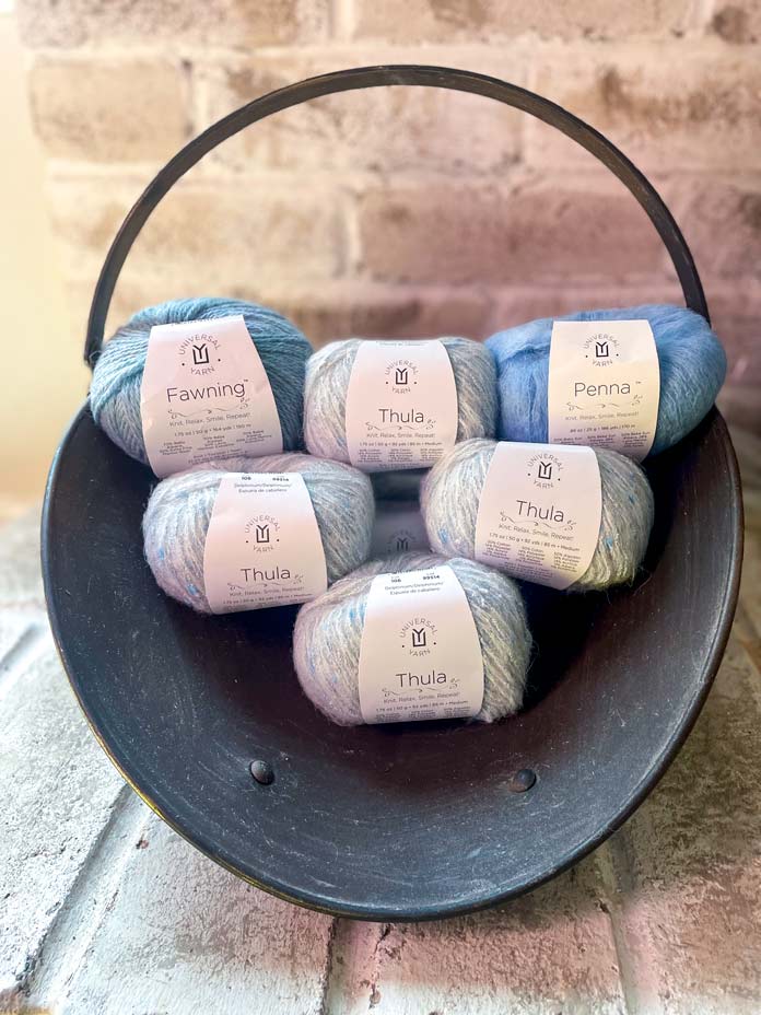 Five balls of UNIVERSAL YARN Thula in the Delphinium colorway, one ball of UNIVERSAL YARN Fawning in the Moonstone colorway, and one ball of UNIVERSAL YARN Penna in the Clouded colorway in a firewood holder on bricks.