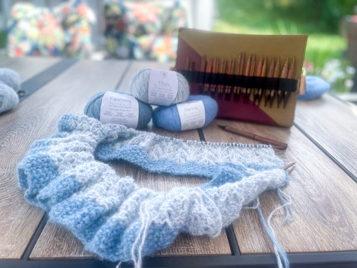 A knitted portion of Lace Tipped Cardigan, one ball of UNIVERSAL YARN Thula in the Delphinium colorway, one ball of UNIVERSAL YARN Fawning in the Moonstone colorway, one ball of UNIVERSAL YARN Penna in the Clouded colorway, a pen, and Knitpro Ginger Special Deluxe Interchangeable Needle Set on a table.