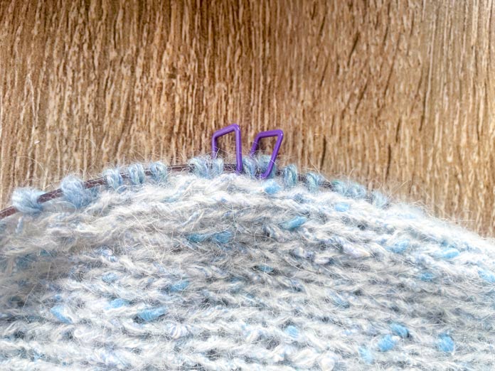A knitted portion of Lace Tipped Cardigan using UNIVERSAL YARN Thula in Delphinium with the first row of blue trim knitted using UNIVERSAL YARN Fawning in Moonstone and UNIVERSAL YARN Penna in Clouded stranded together on a Knitpro Ginger Special Deluxe Interchangeable Needle Set knitting cable. One stitch has markers on each side of it.