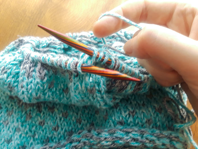A Holland Cowl in progress on a set of Knit Picks Rainbow Wood Circular Needles. It’s knit in two colors of Universal yarns Mystical Marl. Someone is holding two strands of Mystical Marl and twisting them to lock the floats together on the wrong side of the work.