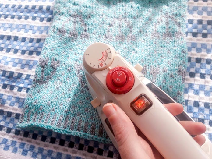 A finished Holland Cowl knit in UNIVERSAL YARNs Mystical Marl - an acrylic yarn blend. The finished cowl is laid flat and being pressed with a steam iron.