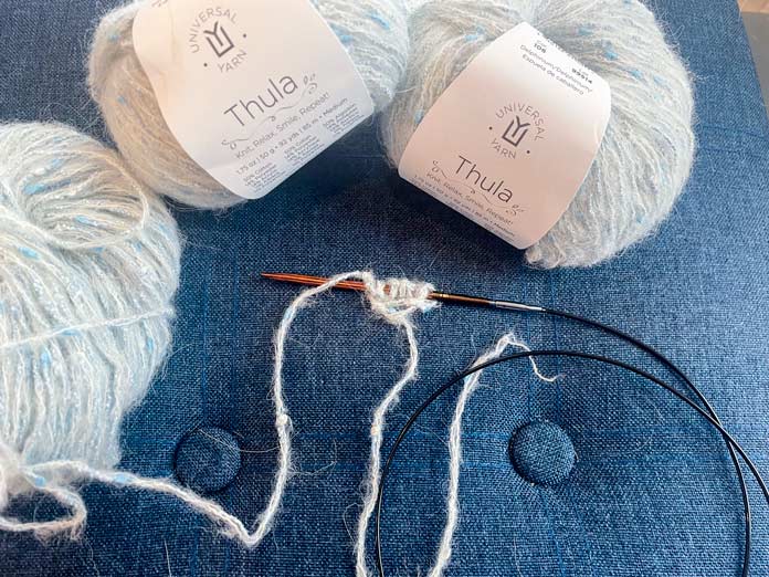 3 balls of yarn and a knitted row on a knitting needle on a blue surface. 