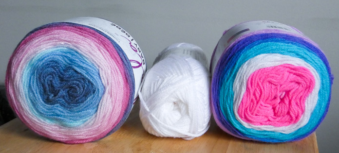 3 balls of yarn sit on a wooden tray. The ball on the left, Arendelle, has denim blue in the center, ringed by light blue, pale blue, light pink, pink, medium rose, and denim. A ball of solid white is in between it and a ball of Unicorn Cloud, which has hot pink in the center, ringed with white, bright blue, medium blue, dark blue and lilac.