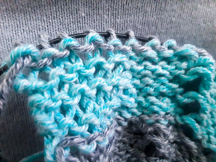 The Wrong Side of the Sunshine Throw Worsted Blanket on the needles showing where the purl stitches distort the lines of the stripes
