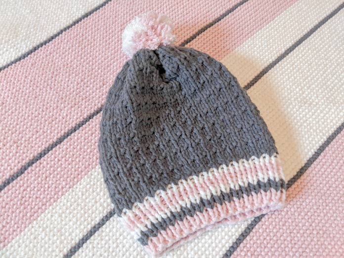 The PomPom Baby Hat knit in gray with a white & pink brim and pompom sits on a striped pink, white, and gray knit blanket.