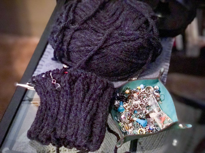 On a glass table a partially knit ribbed brim of a gray hat sits on a circular set of knitting needles beside a tray of stitch markers.