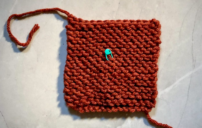 The first square of Rockwell Blanket in the rust color Spicy is displayed with the bind off tail to the top left and a green locking stitch marker placed in the center of the front; Clover Locking Stitch Markers, Hue+Me yarn, Lion Brand Yarn