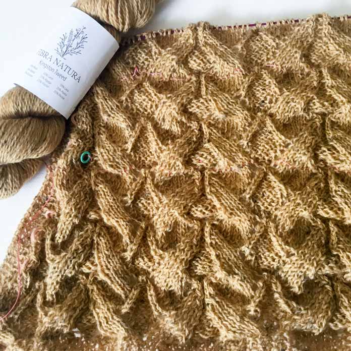 The progressing Arum Shawl with decreases on one side and a lifeline next to a skein of Fibra Natura Kingston Tweed - Ochre