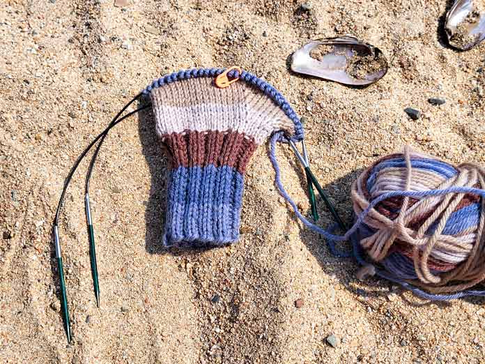 Laying on the sand of a beach is a blue and brown striped mitt with the thumb gusset completed and the yarn attached to the ball, worked in UNIVERSAL YARN Deluxe Stripes, color Timber, knit on 2 KNITTER’S PRIDE Dreamz circular needles.