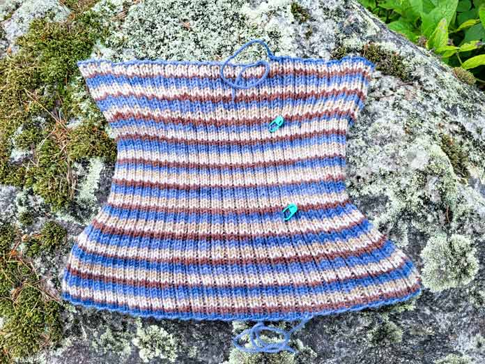 A blue and brown striped cowl laying on a mossy rock shows the curved shape before blocking, knit from UNIVERSAL YARN Deluxe Stripes, color Timber