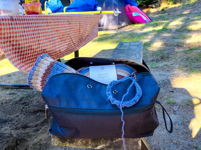 Sitting at a picnic table with a red tablecloth, the second boot cuff is started to be knit on KNITTER’S PRIDE Dreamz circular needles, with the open black VIVACE Knitting Bag showing a ball of UNIVERSAL YARN Deluxe Stripes, color Timber, and a completed blue and brown striped boot cuff. 