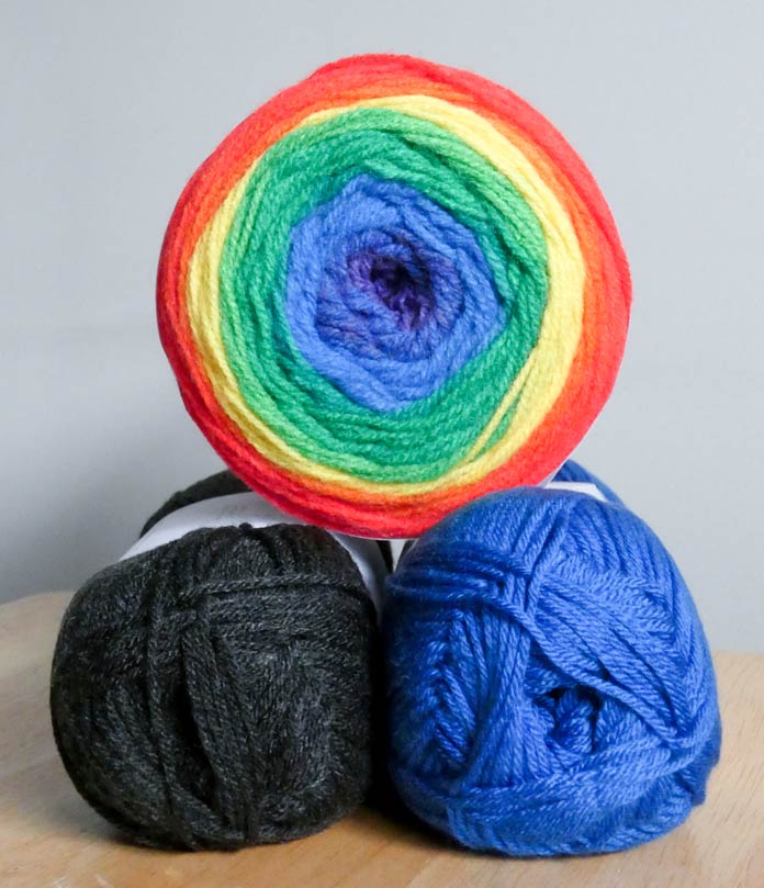 A ball of Rainbow Falls yarn with dark blue in the center, surrounded by rings of bright blue, green, lime green, yellow, orange, and red sits atop two balls of solid yarn: charcoal gray on the left and bright blue on the right.