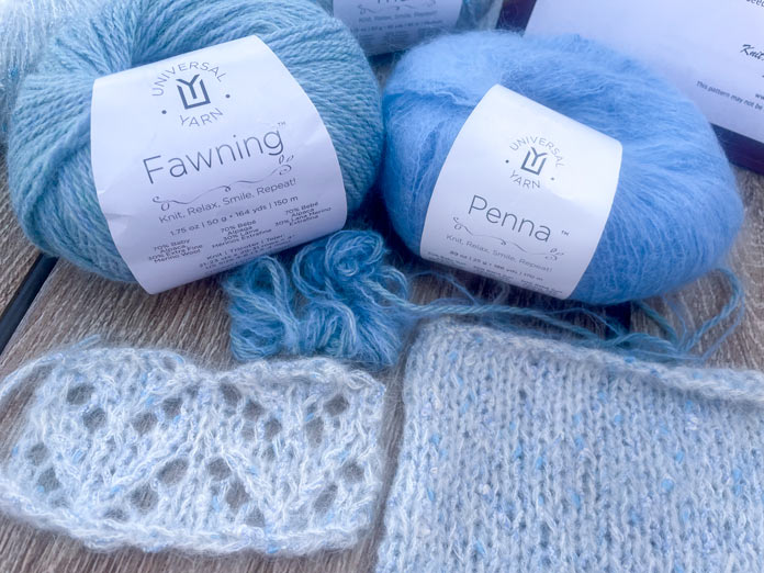 A knitted swatch in lace and a knitted swatch of stockinette in UNIVERSAL YARN Thula in the Delphinium colorway, one ball of UNIVERSAL YARN Fawning in the Moonstone colorway, one ball of UNIVERSAL YARN Penna in the Clouded colorway, and some blue strands of yarn wound together.