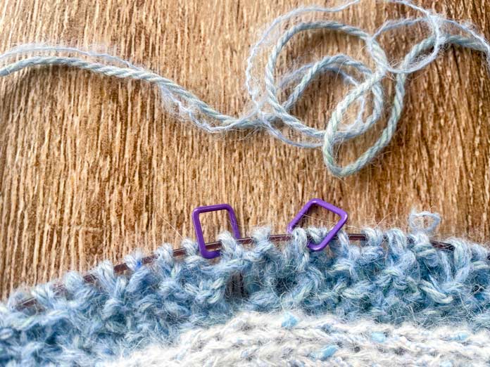 A knitted portion of Lace Tipped Cardigan using UNIVERSAL YARN Thula in Delphinium with the first row of blue trim knitted using UNIVERSAL YARN Fawning in Moonstone and UNIVERSAL YARN Penna in Clouded stranded together on a Knitpro Ginger Special Deluxe Interchangeable Needle Set knitting cable. Three stitches are between knitting markers.