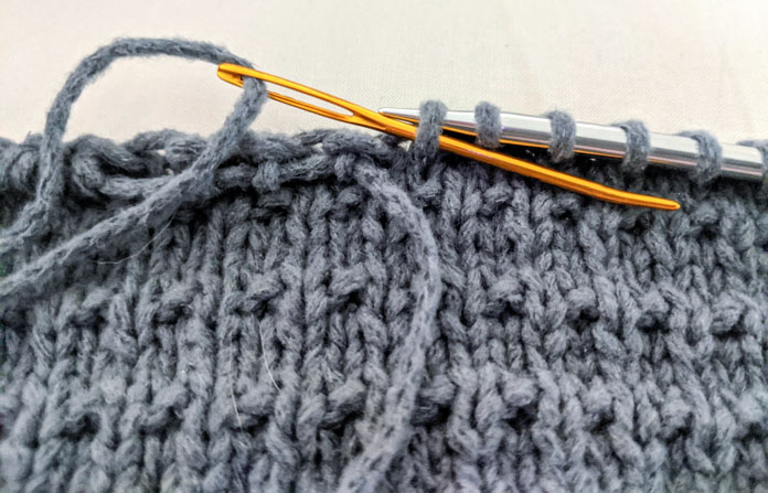 A large gold yarn passes through the gray loops on a metal knitting needle.