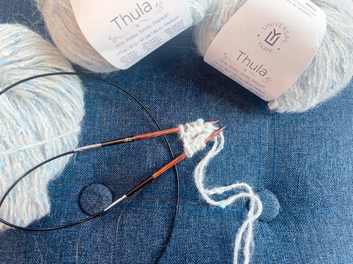 3 balls of yarn and a knitted row on a knitting needle on a blue surface.