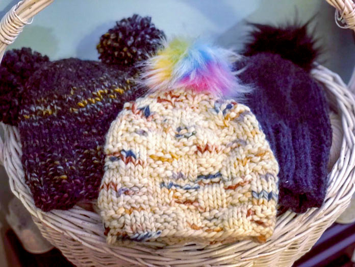 Three different bulky hats with pompoms are nestled in a large white basket.