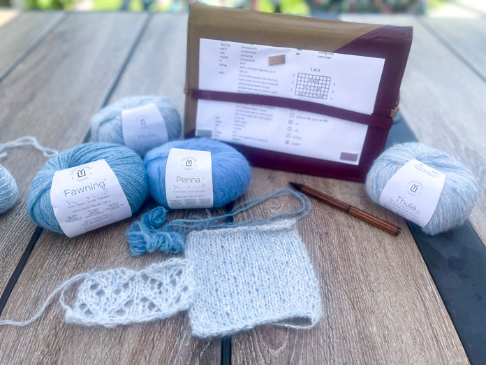 Two balls of UNIVERSAL YARN Thula in Delphinium, one ball of UNIVERSAL YARN Fawning in Moonstone, one ball of UNIVERSAL YARN Penna in Clouded, a stockinette swatch, a lace swatch, and the KnitPro Ginger Special Deluxe Interchangeable Needle Kit Magnetic Pattern Holder hold the Lace Tipped Cardigan pattern.
