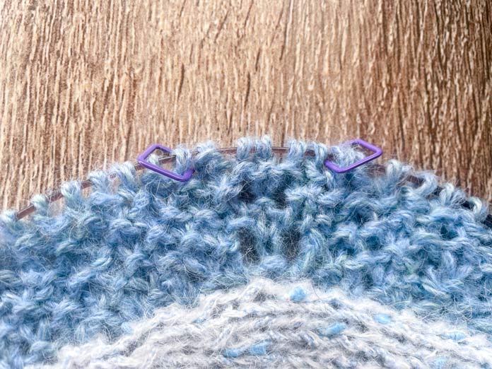 A knitted portion of Lace Tipped Cardigan using UNIVERSAL YARN Thula in Delphinium with the first row of blue trim knitted using UNIVERSAL YARN Fawning in Moonstone and UNIVERSAL YARN Penna in Clouded stranded together on a Knitpro Ginger Special Deluxe Interchangeable Needle Set knitting cable. Five stitches are between knitting markers.