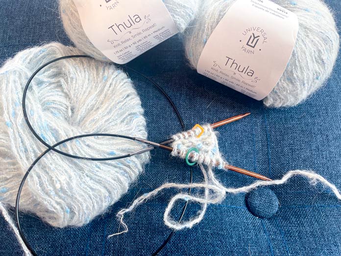 3 balls of yarn and knitted rows on a knitting needle on a blue surface.