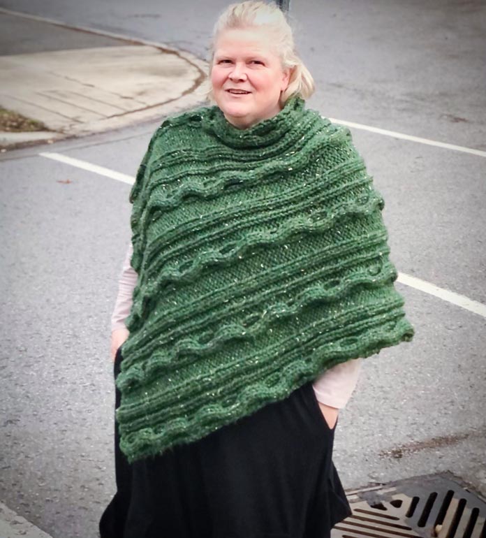 Fiona stands out on the sidewalk wearing her green cabled poncho