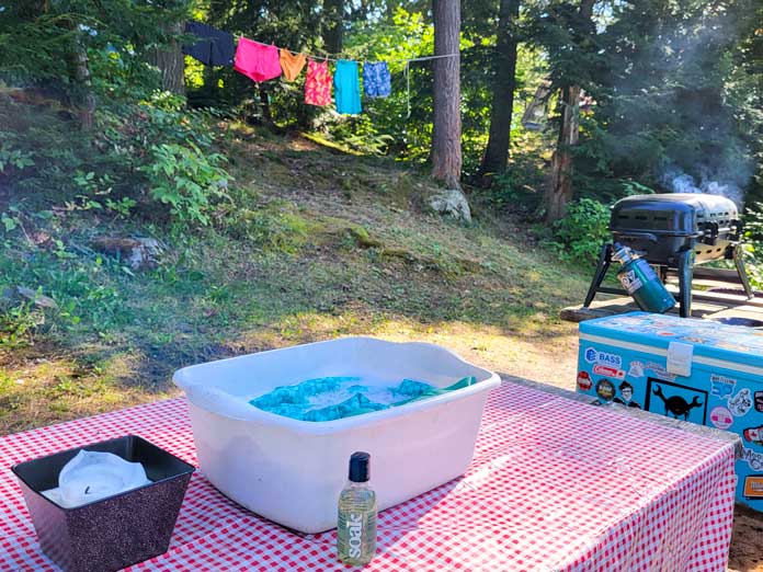 A clothesline with colorful swimsuits hangs in the background of a red table-clothed picnic table where a bottle of Soak gentle laundry soap sits in front of a whitewash basin holding water and blue swim trunks; a candle, a smoking BBQ, and a blue cooler covered in stickers is also visible.