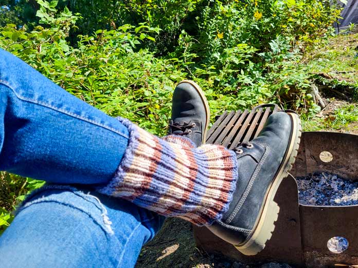 A picture of crossed legs in denim jeans and black hiking boots wearing the blue and brown striped boot cuffs knit from UNIVERSAL YARN Deluxe Stripes, color Timber, feet propped up onto a metal fireplace. 