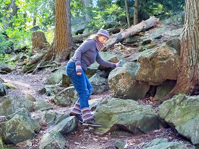 Michelle Porter is stepping down from a rock while hiking, wearing a blue and brown striped hat, mitts, cowl, and boot cuffs, knit from UNIVERSAL YARN Deluxe Stripes, color Timber.