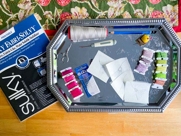 A package of Sulky Sticky Fabri-Solvy near a tray with DMC cotton floss, DMC Chenille needles and sewing notions; Sulky Sticky Fabri-Solvy, DMC floss, DMC Chenille needles