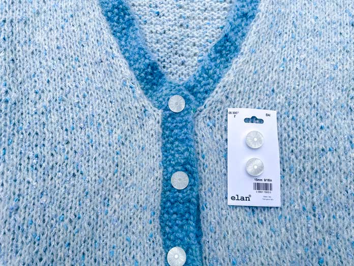 A knitted neck of Lace Tipped Cardigan using UNIVERSAL YARN Thula in Delphinium with blue trim knitted using UNIVERSAL YARN Fawning in Moonstone and UNIVERSAL YARN Penna in Clouded stranded together and Elan 15 mm white iridescent buttons