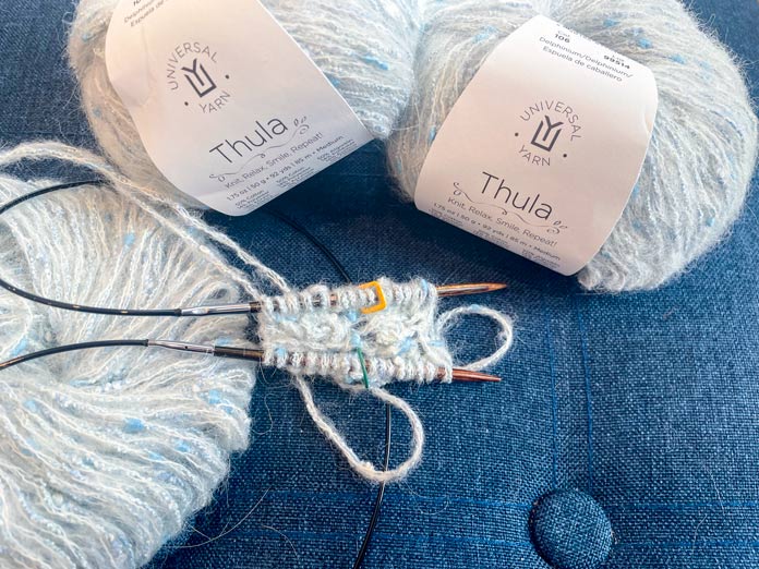 3 balls of yarn and knitted rows on a knitting needle on a blue surface.