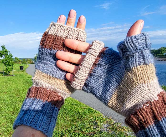 : Two upheld hands wearing blue and brown striped open finger mitts knit from UNIVERSAL YARN Deluxe Stripes, color Timber, showing one with folded ribbing, against a background of a walking trail along a river.